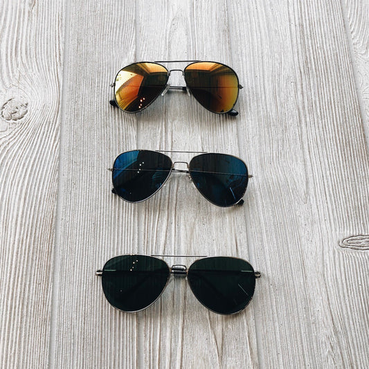 Three pairs of Mac Sunglasses • Adult with UV400 protection—featuring gold, blue, and black lenses—are neatly arranged vertically on a light-colored wooden surface.