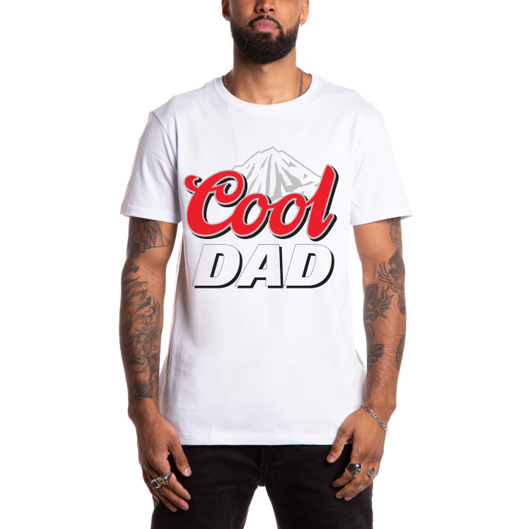 A man wears the Cool Dad • White Tee, a soft unisex shirt made from combed ring-spun cotton, featuring bold red and black Cool DAD text with a mountain illustration in the background.