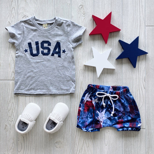 Flat lay featuring the USA • Gray Kids Tee paired with blue-red-white tie-dye shorts, white shoes, and red, white, and blue star décor—creating a fun patriotic look on a light wood background.