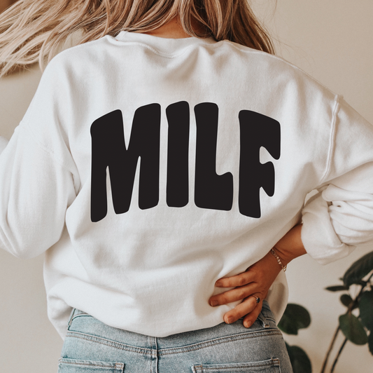 A woman with long blonde hair, facing away from the camera with one hand on her hip, models the MILF • White Sweatshirt— a unisex-fit white pullover featuring MILF in bold black letters on the back.