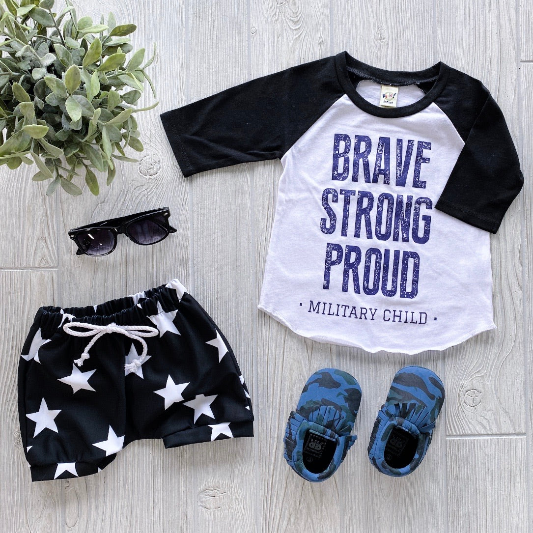 A flat lay showcases the Military Kid Raglan—a white and black shirt with “Brave Strong Proud Military Kid” in blue—paired with black star shorts, blue camo shoes, sunglasses, and a green plant on a light wooden floor.