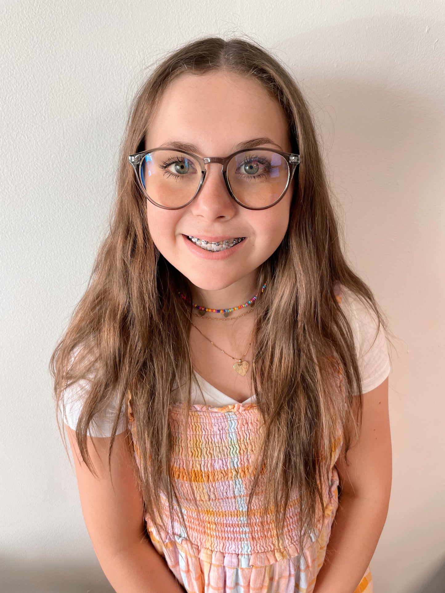 A smiling girl with long brown hair wears Temp Blue Light Glasses in blue and a pastel dress over a white shirt, accessorized with colorful bead necklaces—these glasses help reduce eye strain and headaches. Available in more colors!.