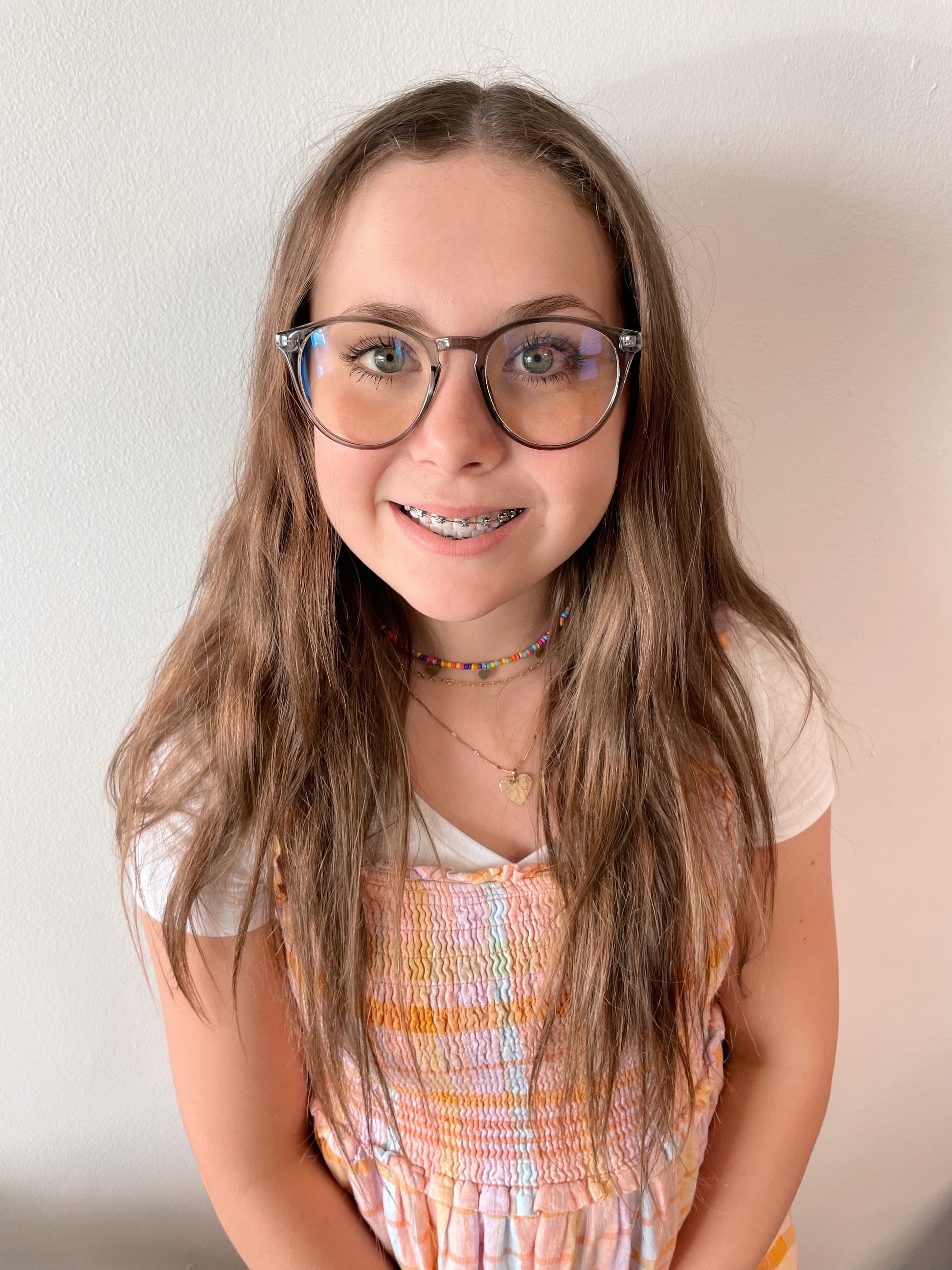 A smiling girl with long brown hair wears Temp Blue Light Glasses in blue and a pastel dress over a white shirt, accessorized with colorful bead necklaces—these glasses help reduce eye strain and headaches. Available in more colors!.