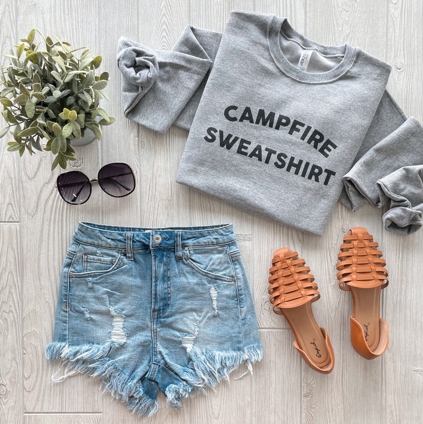A flat lay of a casual outfit featuring the Campfire Sweatshirt • Heather Pullover with unisex fit, distressed denim shorts, tan woven sandals, sunglasses, and a small potted plant on a light wood floor.