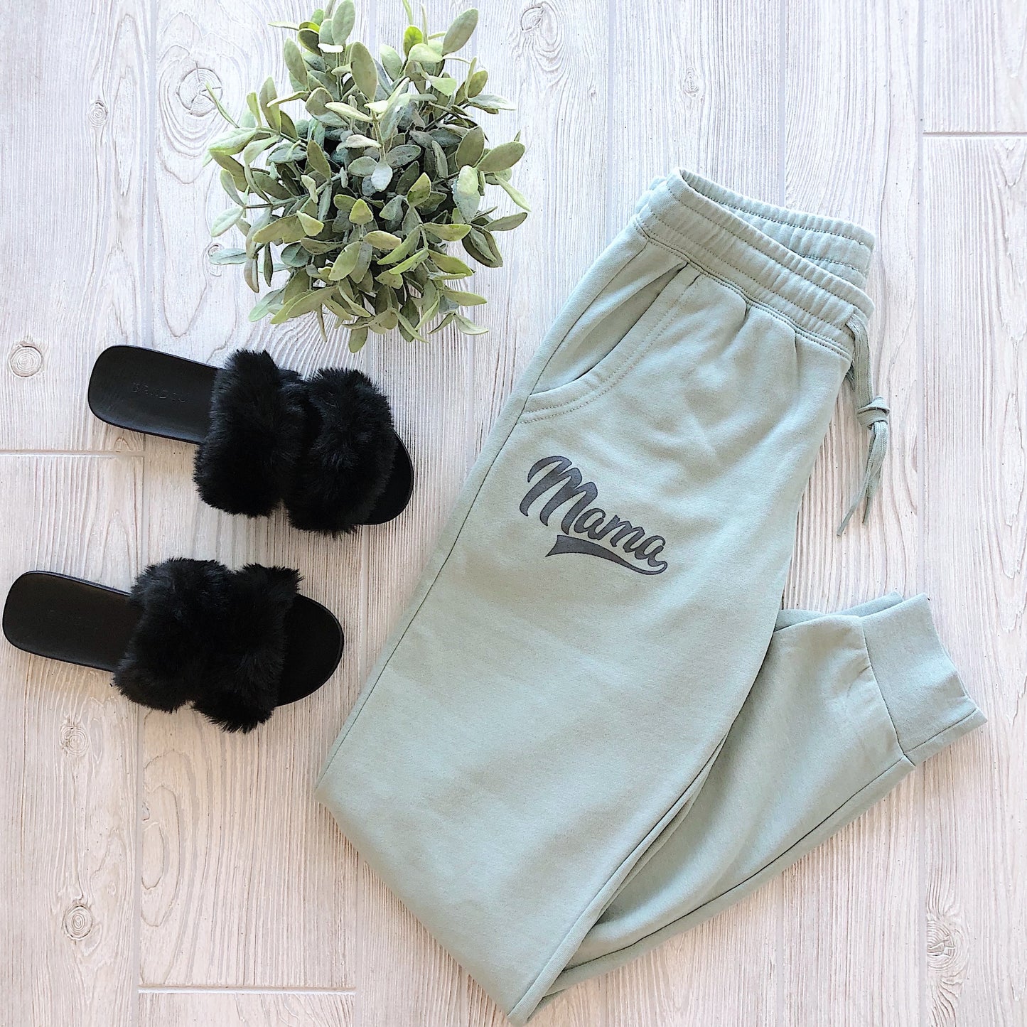 The Mama Jogger • Sage, made from a soft cotton polyester blend, is displayed next to black fuzzy slippers and a small potted plant on a light wooden floor.