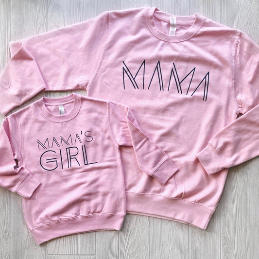 Two pink Mama Line Print• Pink Pullovers on a light wooden floor, made from pre-shrunk cotton/polyester. The larger reads MAMA, the smaller MAMAS GIRL. Both feature a unisex fit and bold geometric font style.