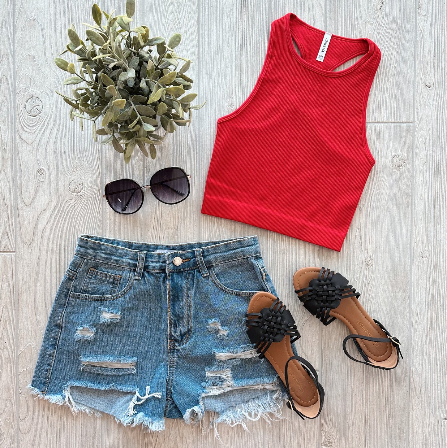 A flat lay of summer clothing: the Sophia High-Neck Crop • Ruby, distressed denim shorts, black woven-strap sandals, black sunglasses, and a small green potted plant on a light wooden surface.