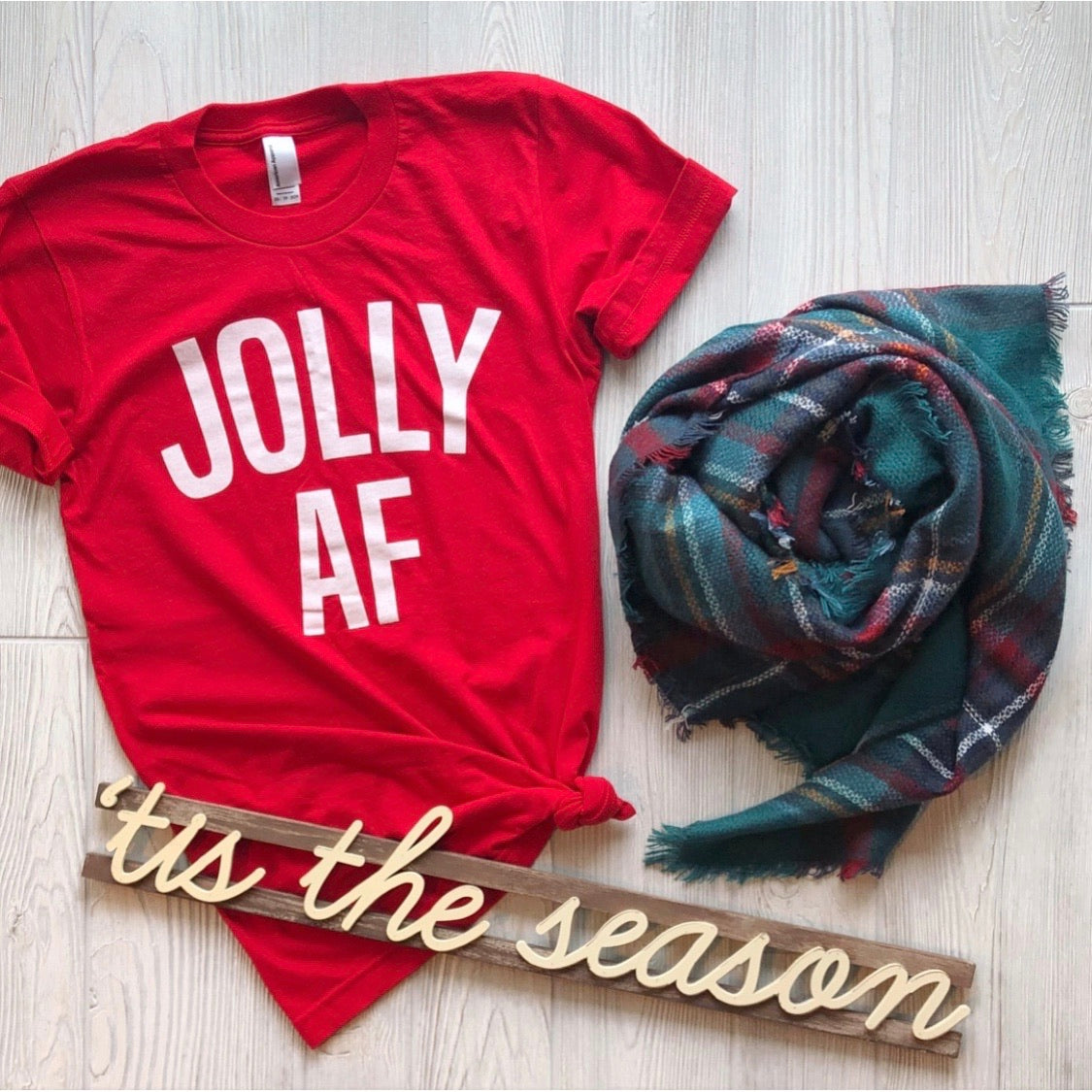 The JOLLY (and fun) • Tee, a unisex red shirt with bold white letters, is styled with a plaid scarf and a festive "tis the season" wooden sign on a light wood floor—ideal for your Christmas shirt collection.