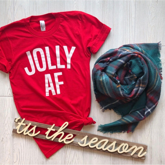 The JOLLY (and fun) • Tee, a unisex red shirt with bold white letters, is styled with a plaid scarf and a festive "tis the season" wooden sign on a light wood floor—ideal for your Christmas shirt collection.