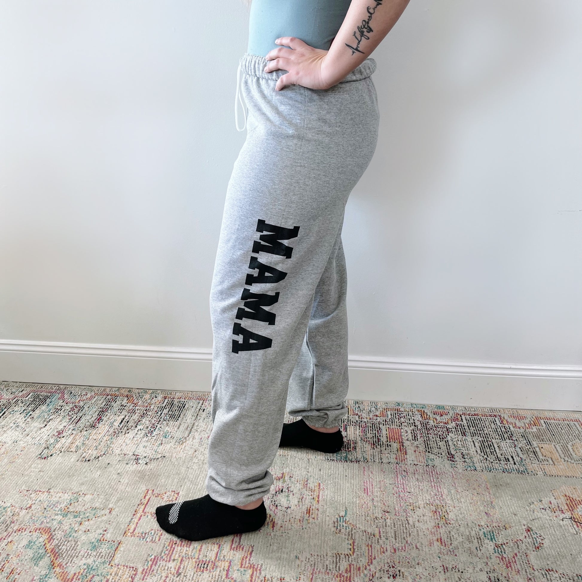 A person stands sideways on a patterned rug wearing MAMA Sweats • gray with bold black lettering, black socks, and a light top. A tattoo is visible on one arm—these cozy sweats are perfect loungewear for relaxing at home.