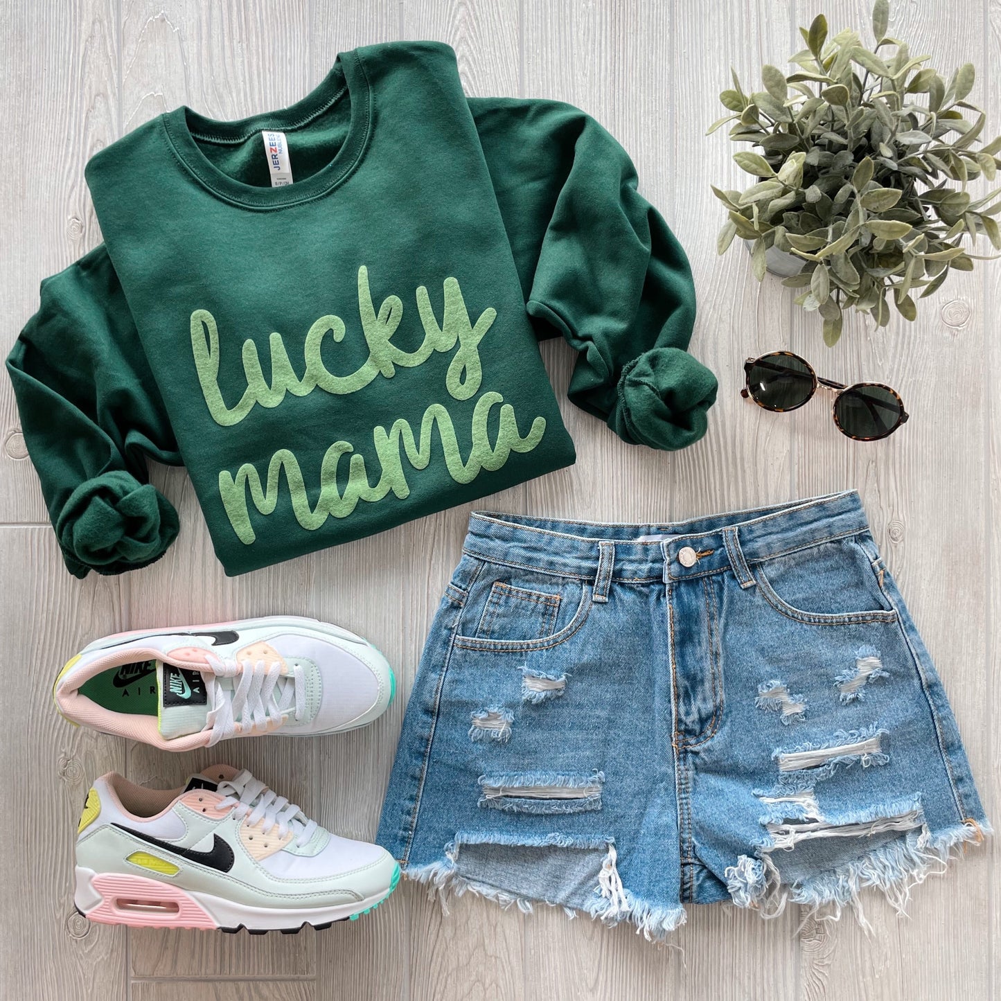 A flat lay features the lucky mama • Forest Pullover in a unisex fit, paired with distressed denim shorts, pastel Nike sneakers, black sunglasses, and an artificial potted plant on a light wood background.