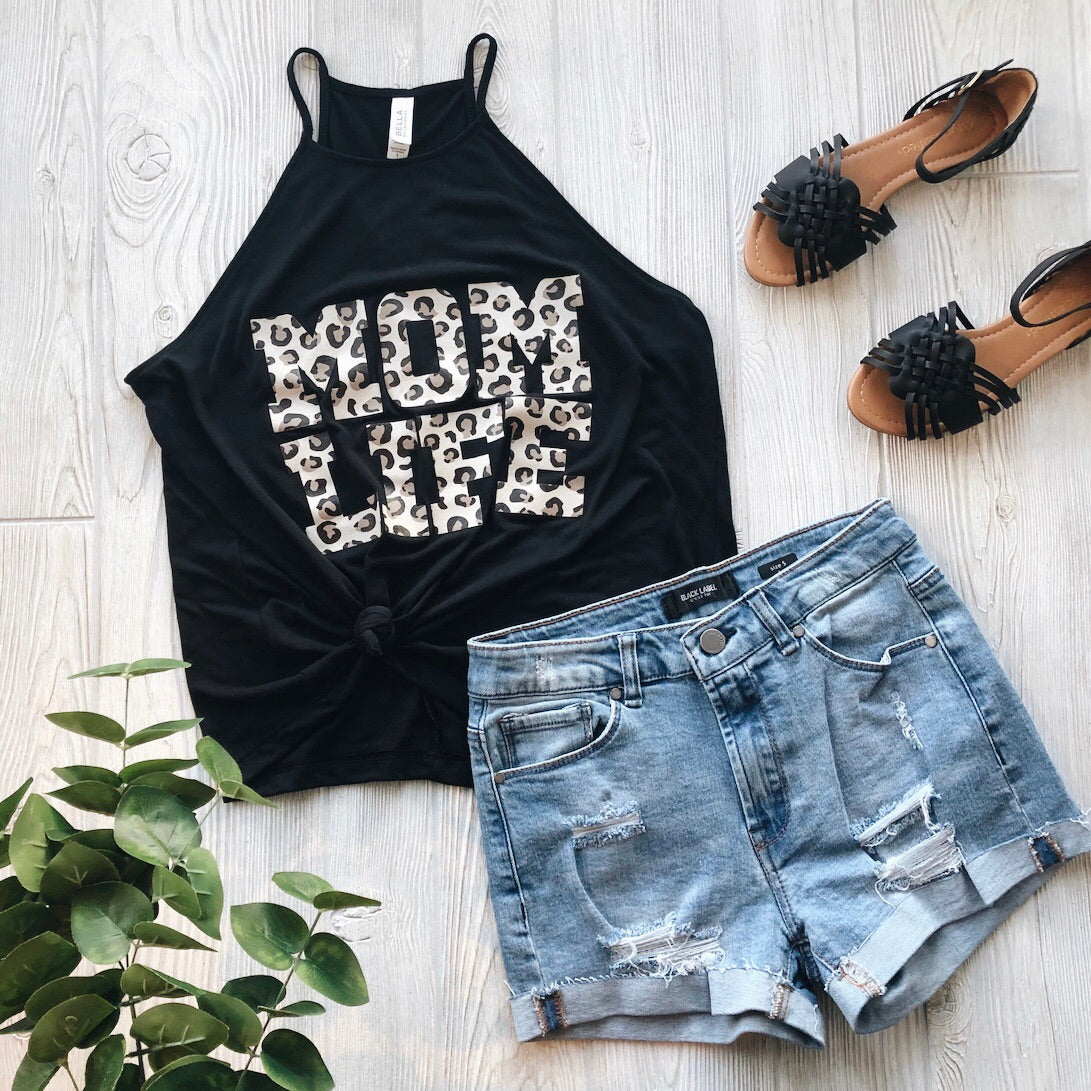 Flat lay of the Mom Life Cheetah • High Neck Tank with a black high neck, paired with distressed denim shorts, black fringe sandals, and a small green plant on a light wood floor.