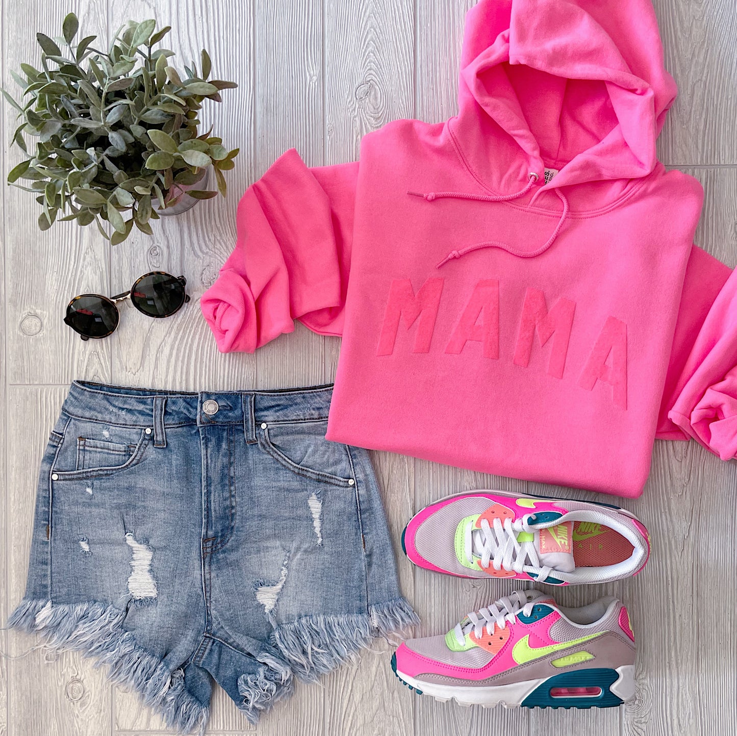 Flat lay of a casual outfit featuring the MAMA arch • Neon Pink Tonal Hoodie in a unisex fit, styled with distressed denim shorts, colorful Nike sneakers, round black sunglasses, and a small green plant on a light wooden floor.