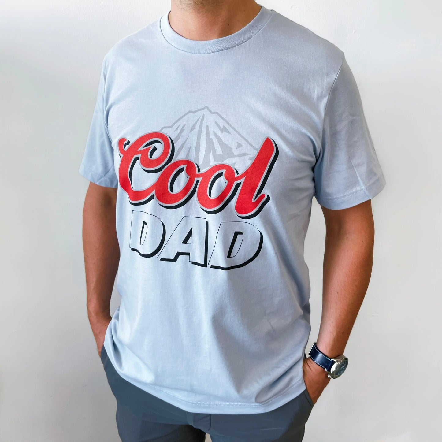 A person wearing the Cool Dad • Ice Blue Tee by Bella Canvas, a unisex t-shirt in light blue featuring bold red and black Cool Dad text with a mountain graphic. Made from soft, combed and ring-spun cotton. Face not visible.
