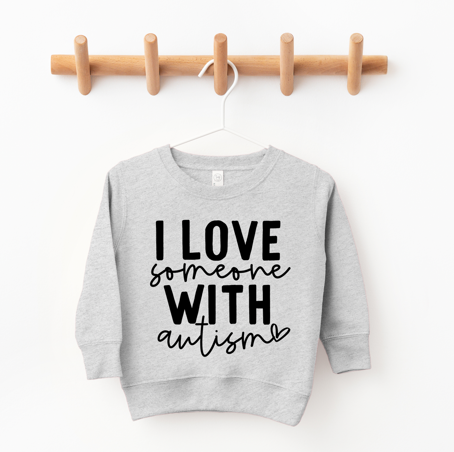 The Autism Love • Kids Pullover, in light gray with bold black I LOVE someone WITH autism text and a small heart, hangs oversized on a wooden peg rack in a childs closet.