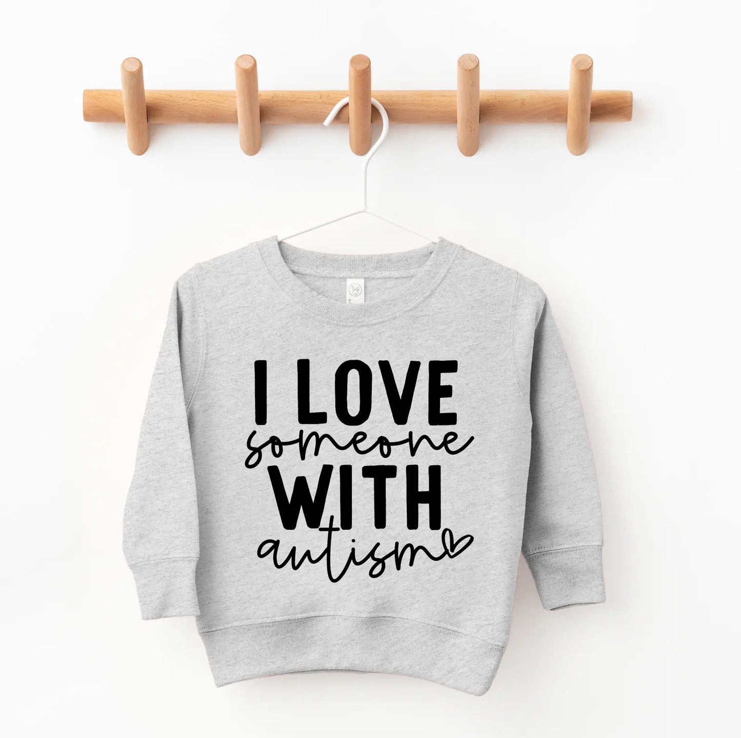 The Autism Love • Kids Pullover, in light gray with bold black I LOVE someone WITH autism text and a small heart, hangs oversized on a wooden peg rack in a childs closet.