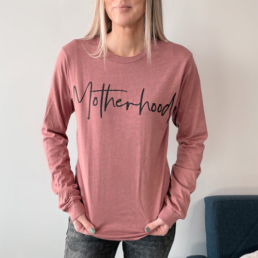 A woman with long blonde hair wears the Motherhood • Mauve Long Sleeve—a wardrobe essential featuring Motherhood in bold black script across the chest. She stands with her hands in jean pockets against a plain background.