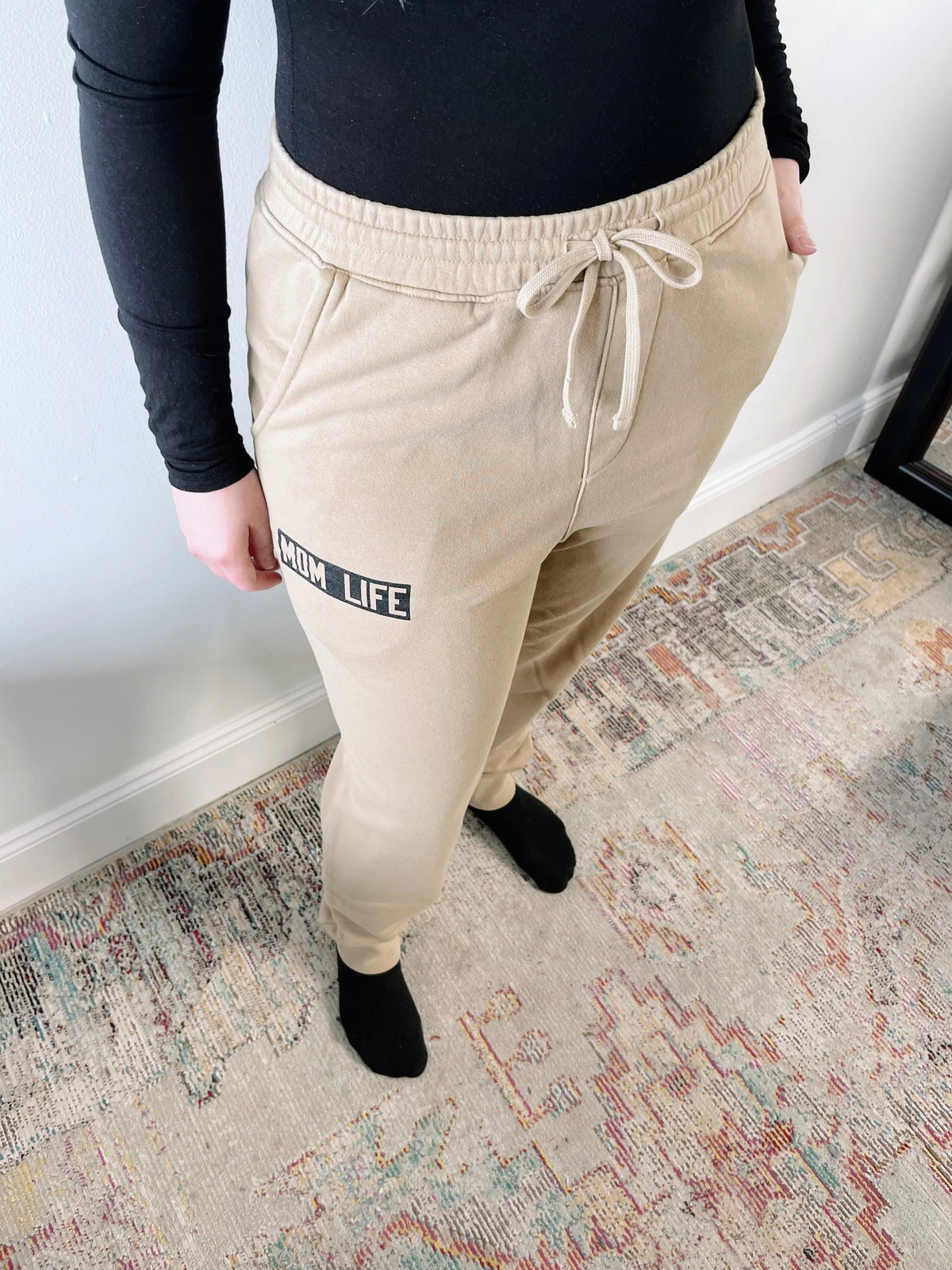 Someone stands on a patterned rug with hands in their pockets, wearing a black long-sleeve shirt and Mom Life Joggers • Natural.