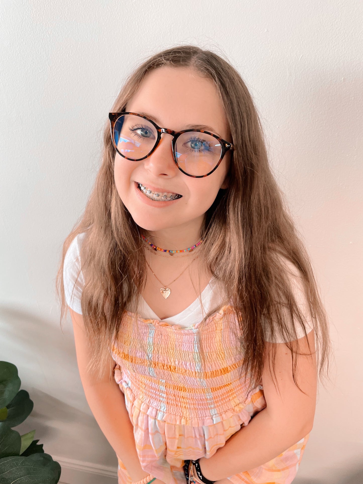 A young person with long brown hair wears Temp • Blue Light Glasses • More Colors!, a white shirt, pastel plaid dress, and colorful choker, standing against a light wall with a green plant to help reduce eye strain.