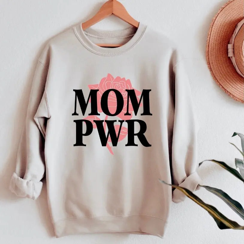 The MOM PWR • Sand Pullover is a light beige unisex sweatshirt with bold black MOM PWR text over a pink rose graphic, displayed on a wooden hanger—ideal for anyone who loves comfy sweatshirts.
