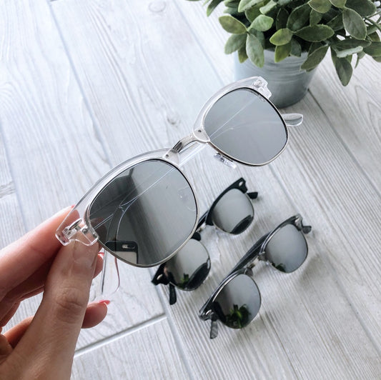 A hand holds Zoe Mirror Sunglasses with transparent frames and reflective lenses above two similar pairs on a light wooden surface; a potted green plant is in the background. Final Sale—Not Eligible for Return. More Colors!.