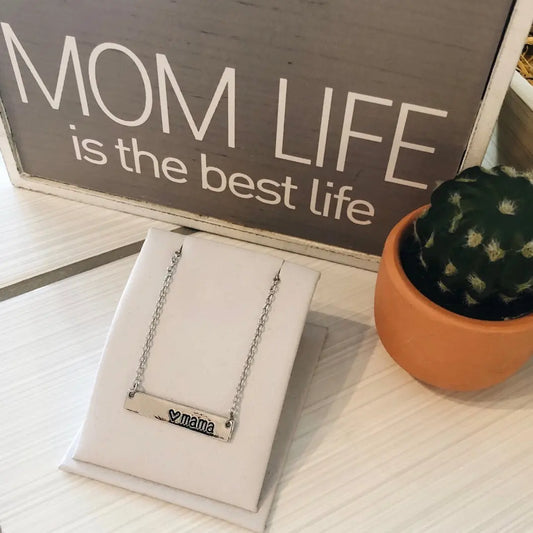 A small cactus in a pot, a gold Mama Heart Necklace, and a sign that reads MOM LIFE is the best life are displayed on a light-colored table.