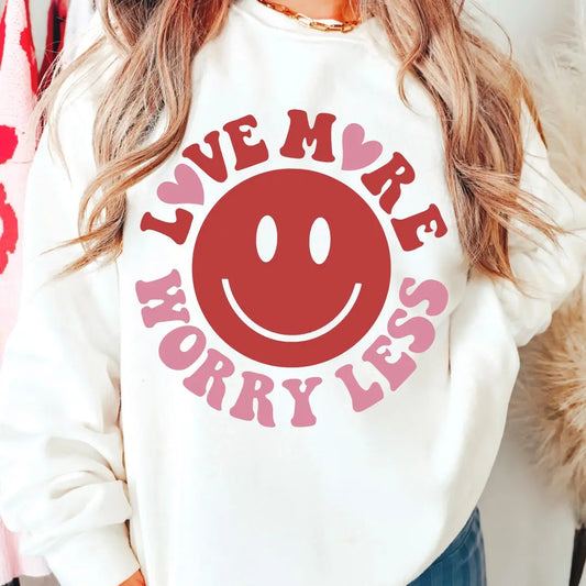 A woman styles the Love More • White Pullover—a cozy unisex fleece with a bold red smiley and pink LOVE MORE WORRY LESS print—pairing its soft cotton/poly blend comfort with long waves and a gold necklace.