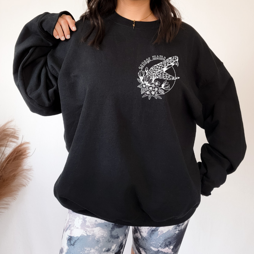 A woman stands against a light background wearing the Savage Mama Snake • Black Pullover featuring a white floral snake design and savage mama on the chest, styled with patterned leggings. This unisex fit offers comfort and bold style.