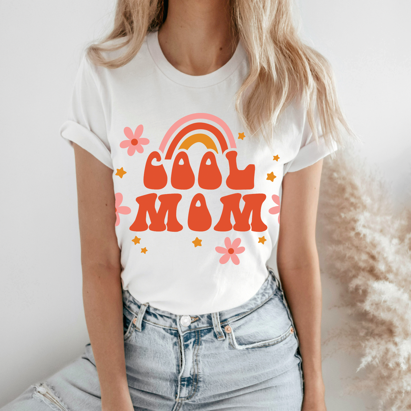 A woman wears the Cool Mom Rainbow • White Tee (unisex fit) with rolled sleeves and light blue jeans. The shirt features retro Cool Mom text, a rainbow, stars, and flowers in orange, pink, and yellow.