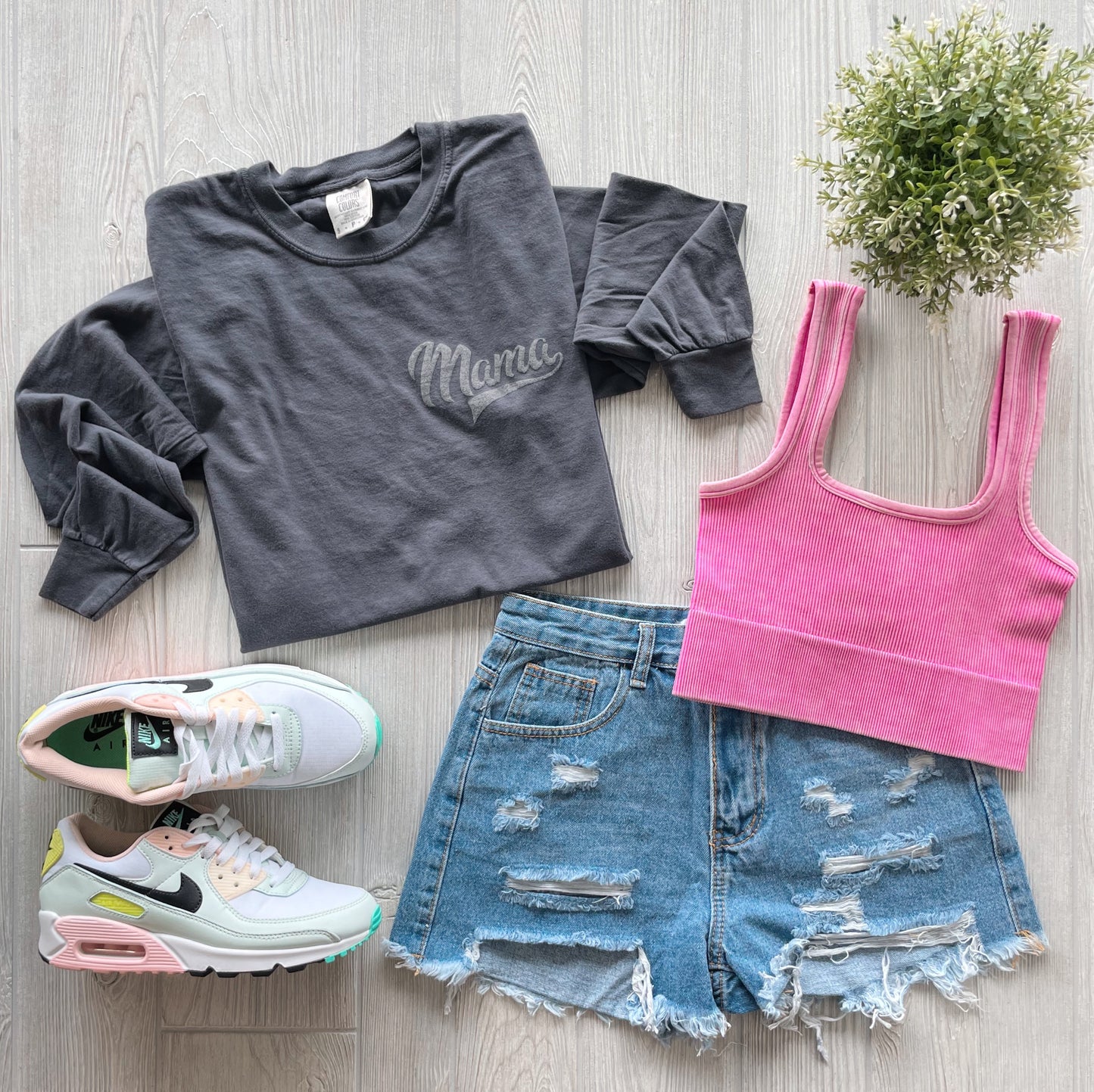 Flat lay of a casual outfit: Mama Sporty • Charcoal Long Sleeve, pink ribbed tank top, distressed denim shorts, white Nike sneakers with pastel accents, and a small green potted plant on a light wood floor.