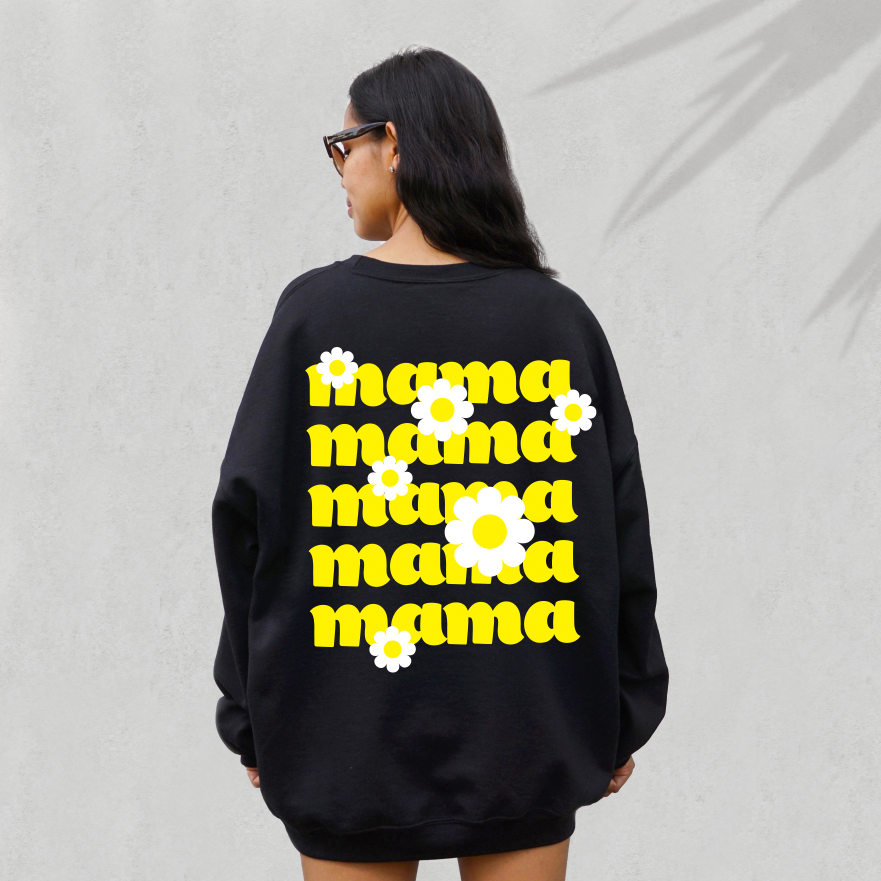 A woman with long dark hair and sunglasses wears the Daisy Mama • Black Pullover, a trendy unisex sweatshirt featuring a bold yellow daisy mama print on the back, as she stands against a light gray wall.