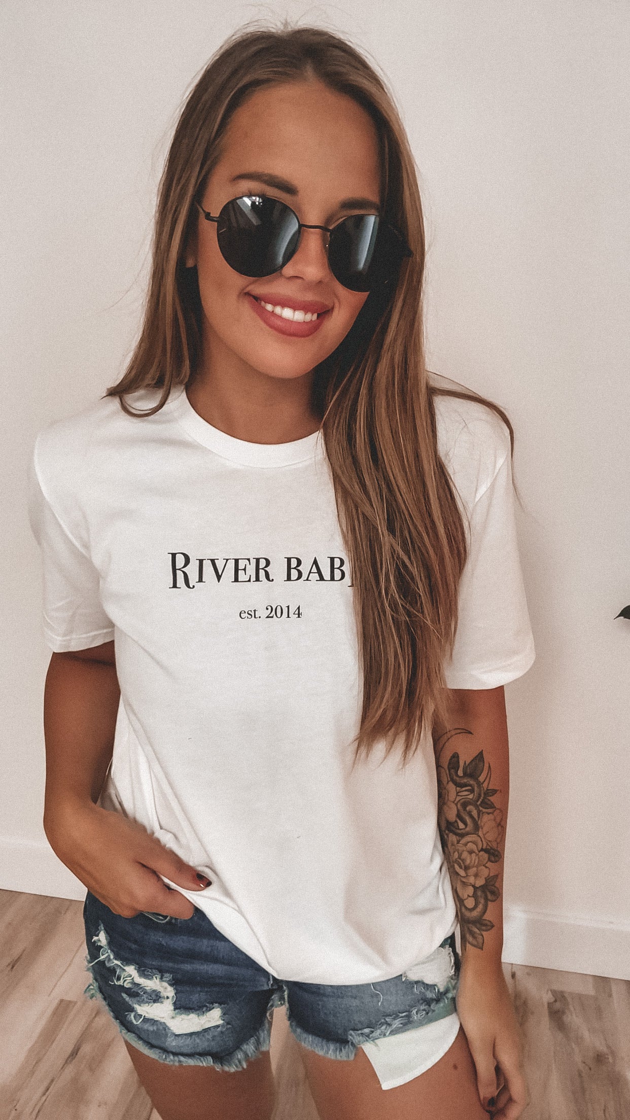 A woman with long brown hair wears Demi • Adult Sunglasses, a white River Babe est. 2014 tee, and ripped denim shorts, smiling against a white wall with a floral tattoo on her arm. Final Sale—Not Eligible for Return.
