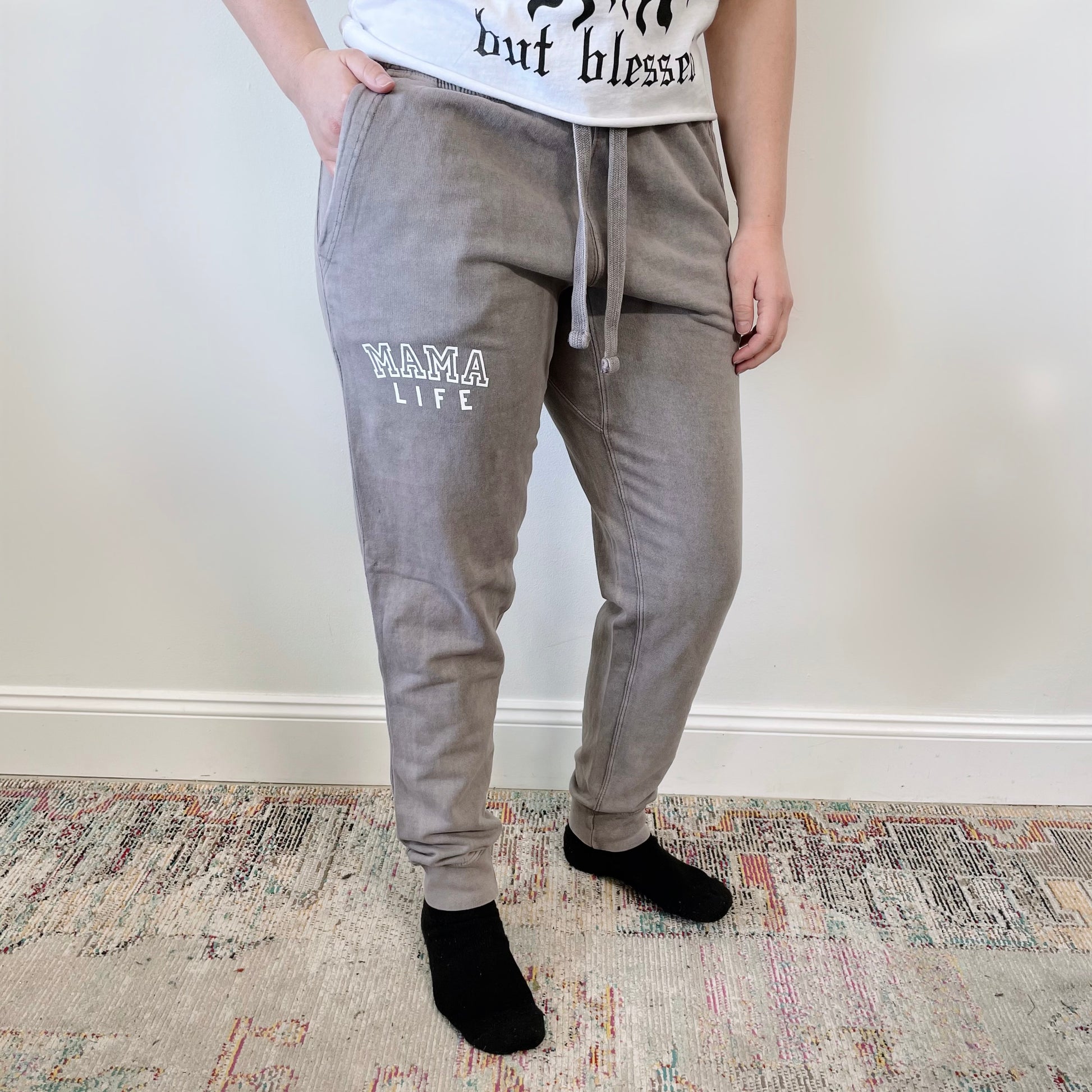 A person stands indoors wearing Mama Life • Acid Wash Joggers in gray, a white graphic t-shirt, and black socks. The background includes a patterned rug and a plain, light-colored wall.