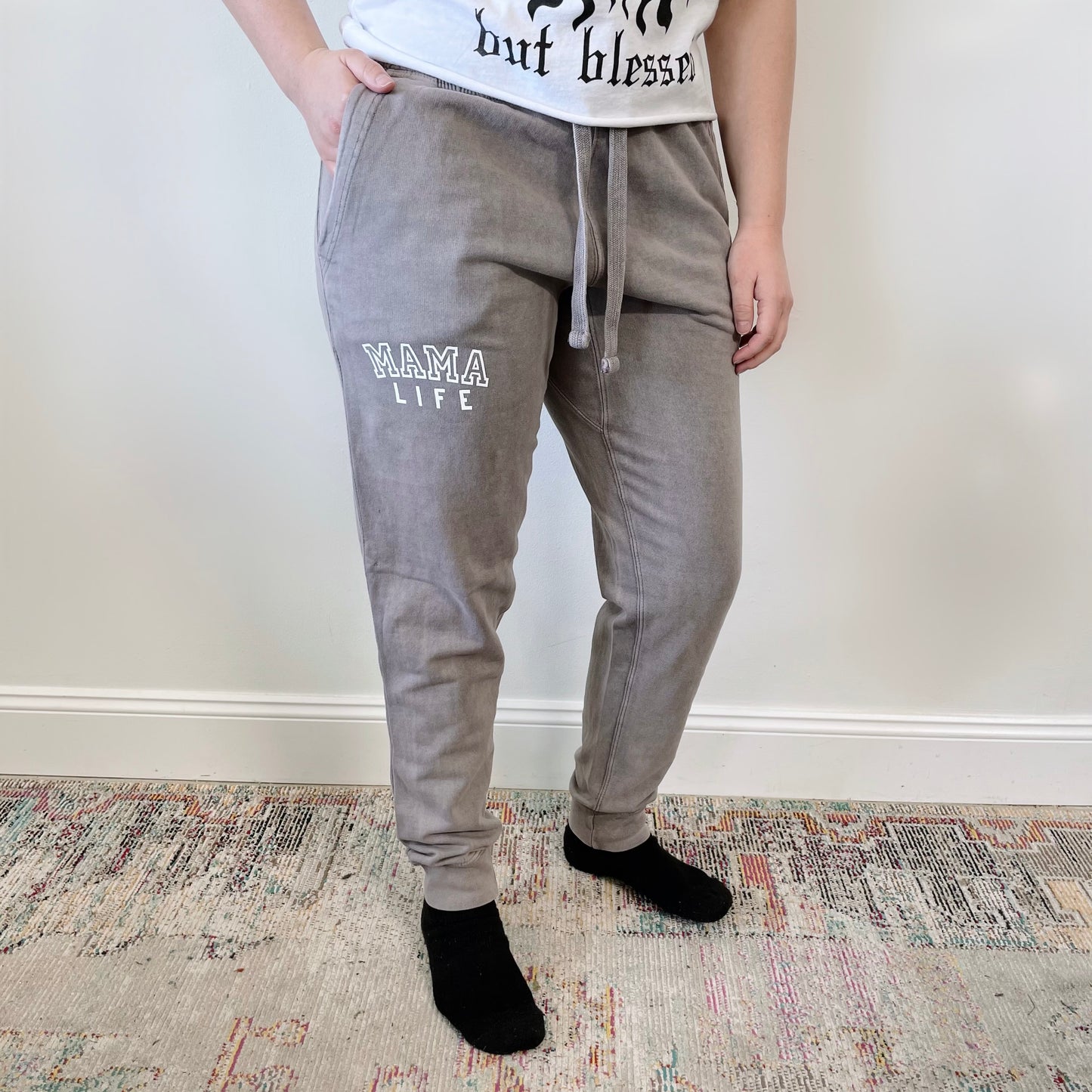 A person stands indoors wearing Mama Life • Acid Wash Joggers in gray, a white graphic t-shirt, and black socks. The background includes a patterned rug and a plain, light-colored wall.
