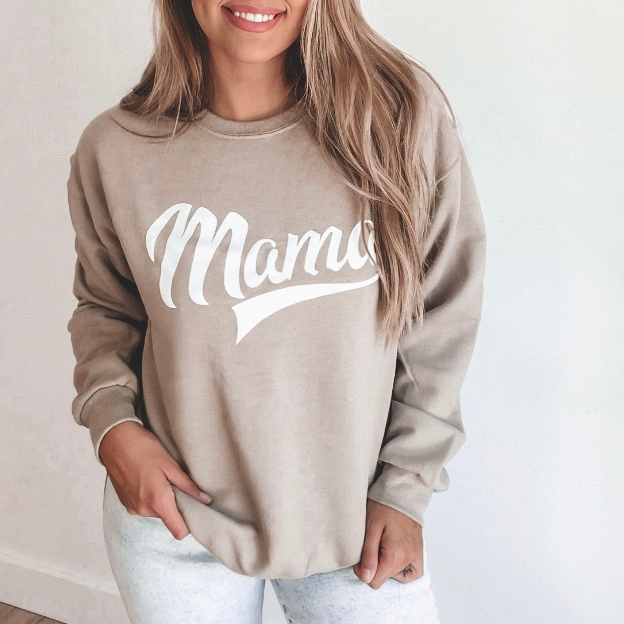 A woman with long, light brown hair smiles in front of a plain wall, wearing the Mama Sporty • Khaki Pullover— a JERZEES unisex fit sweatshirt with “Mama” in white script—paired with light pants.