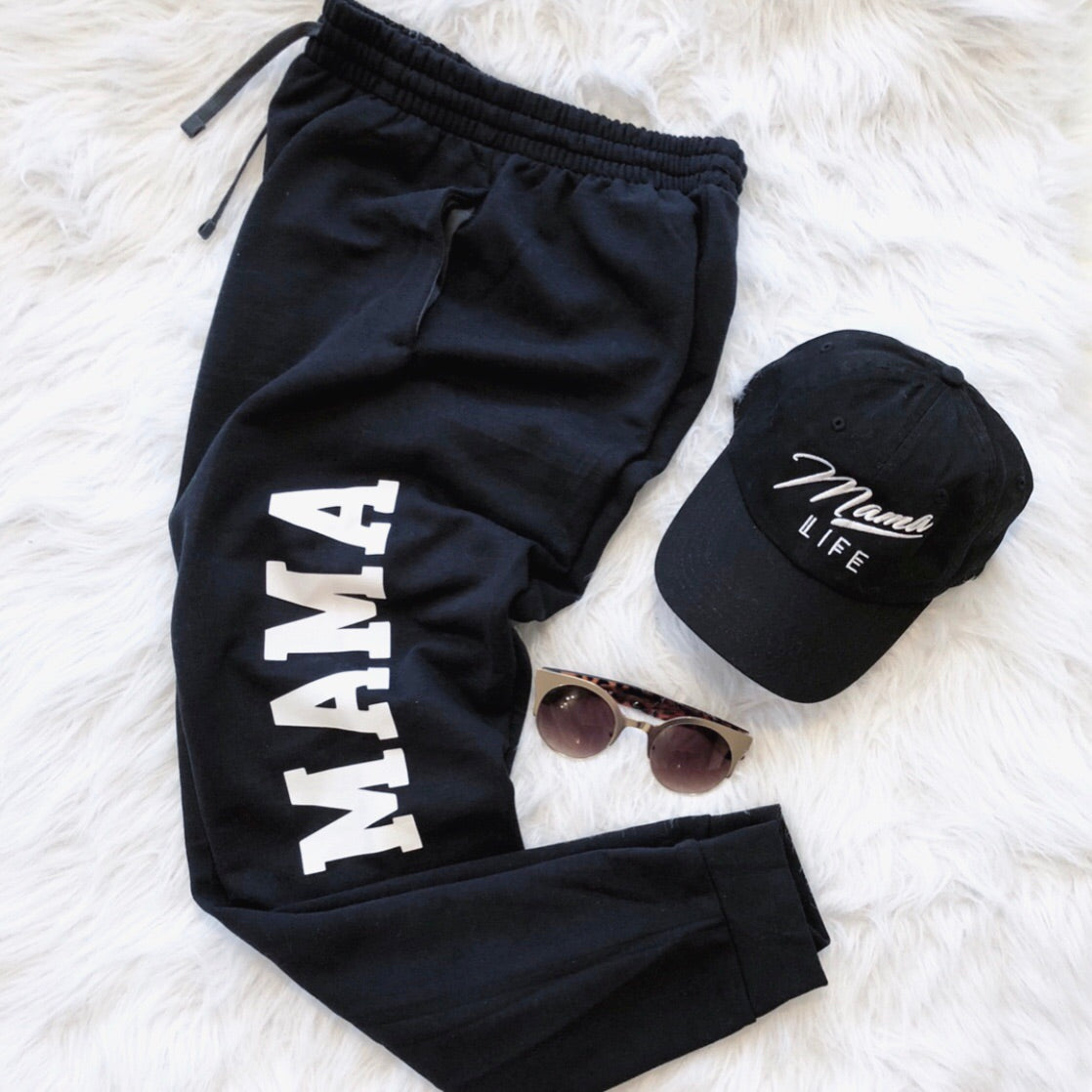 MAMA Joggers in black, paired with a black “Mama Life” cap in white script and brown sunglasses, styled on a soft white surface for the ultimate loungewear look.