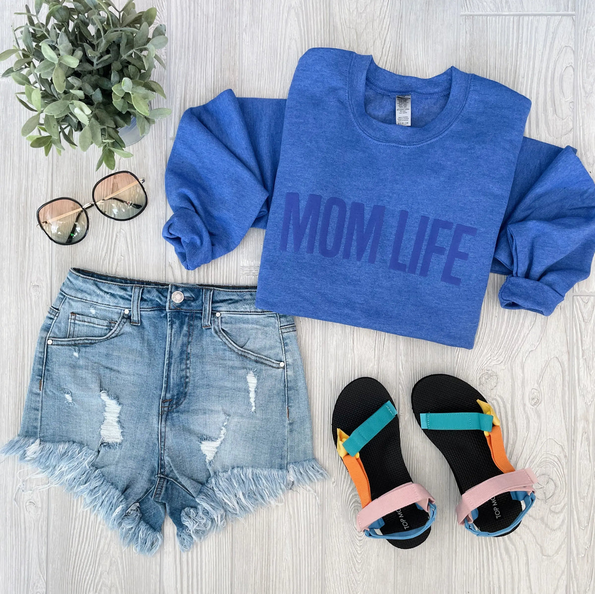 Flat lay of a MOM LIFE • Tonal Periwinkle Pullover, distressed denim shorts, black sandals with colorful straps, sunglasses, and a small potted plant on a light wood surface.