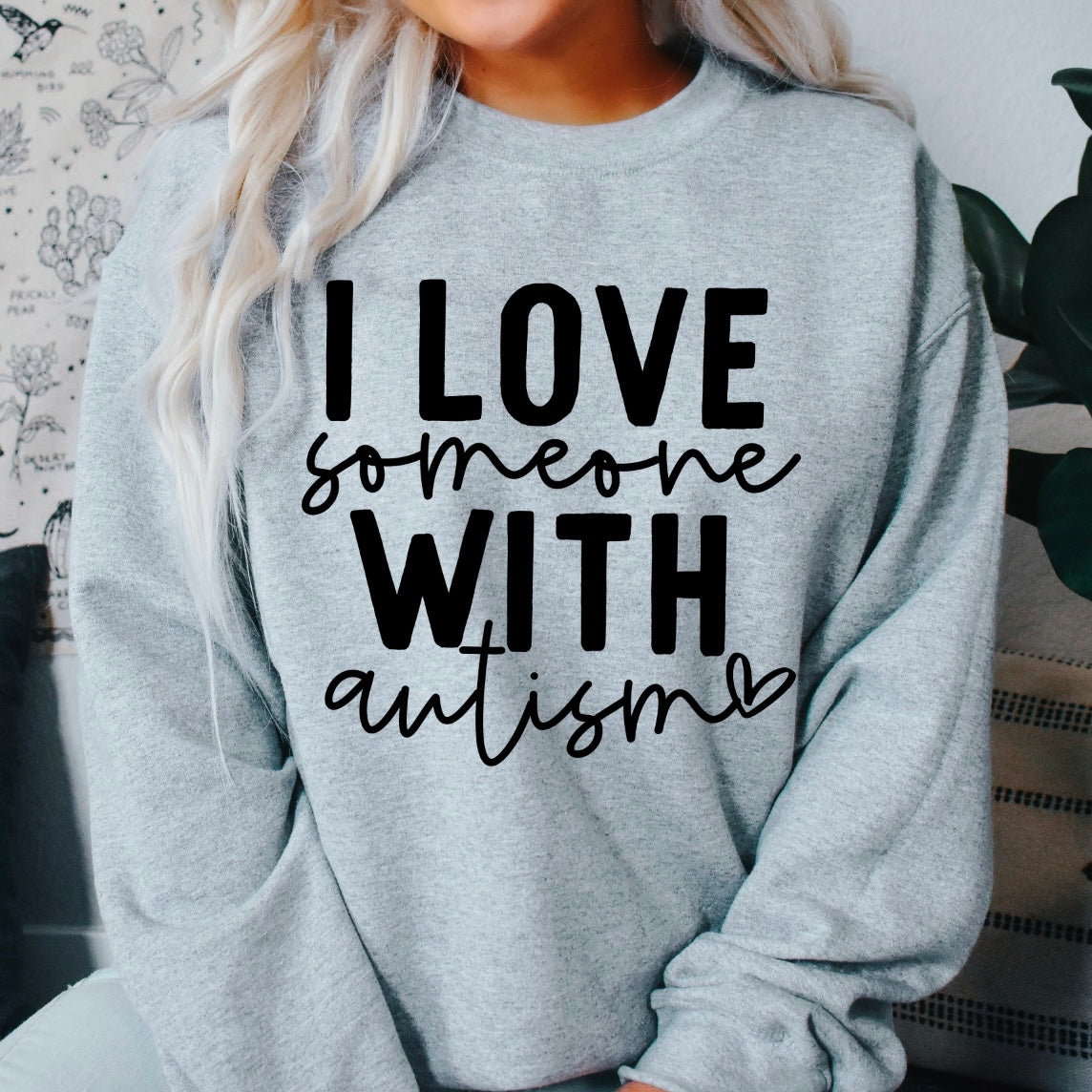 A person in the Autism Love • Gray Pullover, a classic fit crew neck sweatshirt with I LOVE someone WITH autism (script autism and heart) in bold letters—crafted from soft, preshrunk cotton/polyester for lasting comfort.