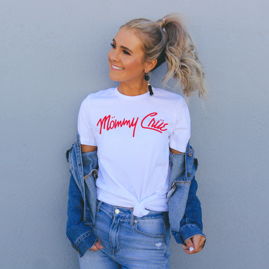 A smiling woman with blonde hair in a ponytail wears a white Mommy Crue tee, blue jeans, and an off-shoulder denim jacket, standing against a light gray textured wall.