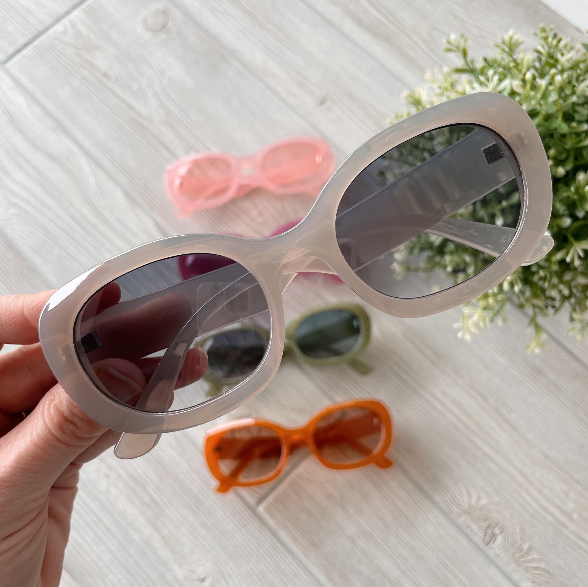 A hand holds Poppy Sunglasses (adult, UV protected) above a light wooden surface. In the background, three more colorful pairs—pink, green, and orange—are arranged near a small potted plant. Final sale item. More colors available!.