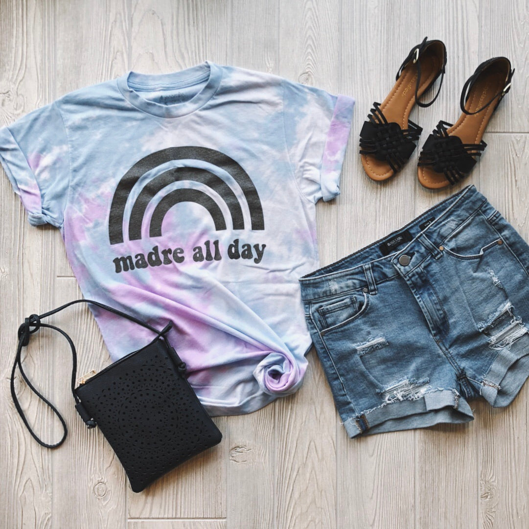 Flat lay of a casual outfit featuring the Madre Rainbow • Purple Tie-Dye t-shirt, distressed denim shorts, black fringe sandals, and a black crossbody purse, all arranged on a light wood floor.