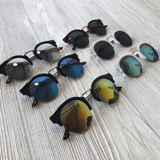 Seven pairs of Ray • Adult Sunglasses with UV400 protection, featuring round lenses in blue, gold, silver, black, and tortoiseshell frames, are displayed on a light wooden surface. Final Sale.