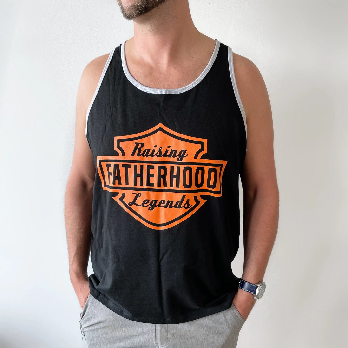 A man wearing the Legends • Ringer Tank—a black Bella/Canvas top with orange Raising Fatherhood Legends text in a motorcycle logo style—stands against a white wall, hands in his pockets.