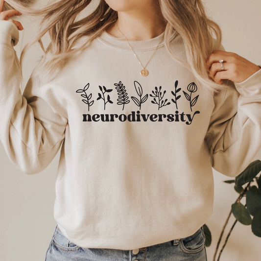 A person stands indoors near a green plant wearing the Neurodiversity • Sand Pullover, a beige fleece-lined unisex sweatshirt with black botanical illustrations and neurodiversity printed on it.