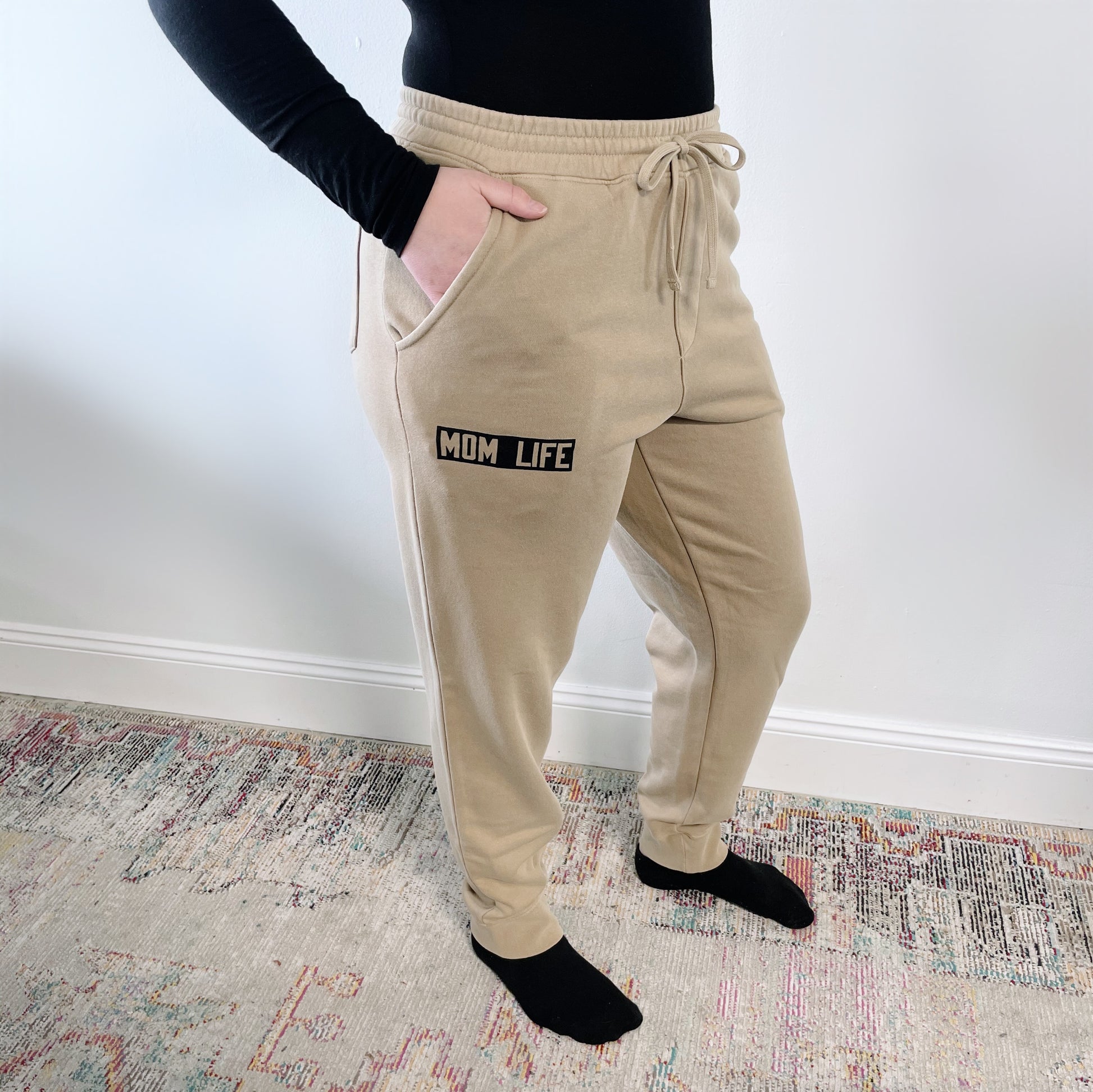 Someone stands on a patterned rug wearing Mom Life Joggers • Natural with MOM LIFE on the left pocket, paired with a black long-sleeve shirt and black socks. Their hand rests in one of the joggers pockets.