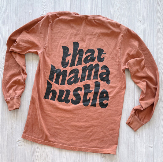 The That Mama Hustle • Rust Comfort Long Sleeve is a soft, garment-dyed shirt with bold black that mama hustle lettering on the back. It’s rust-colored, long-sleeve, and shown laid flat on a light wood surface.