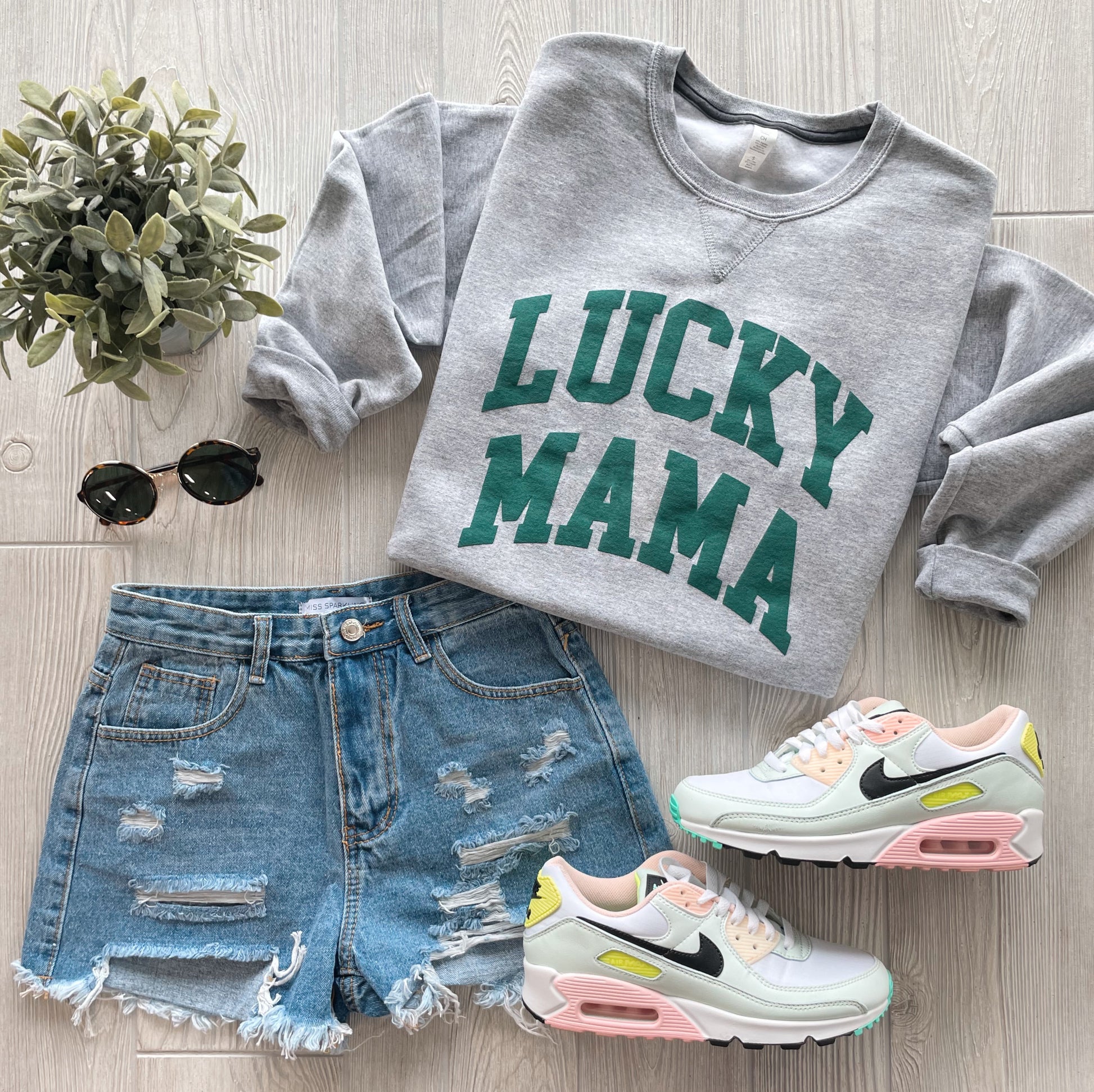 A flat lay of a casual outfit: the LUCKY MAMA • Puff Print Pullover in gray with matte forest text, paired with distressed denim shorts, white pastel-accent sneakers, black sunglasses, and a small potted plant on a light wood floor.