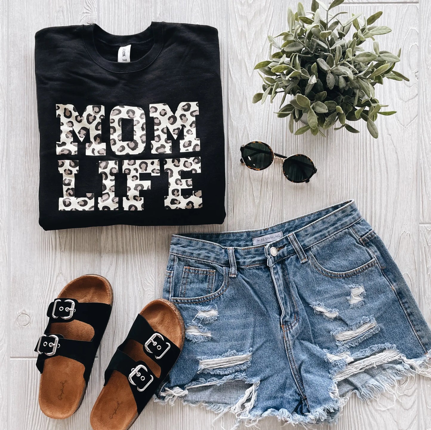 Flat lay of a folded Mom Life Cheetah • Black Pullover with unisex fit, paired with distressed denim shorts, black sandals, round sunglasses, and a small potted plant on a light wooden surface.
