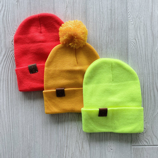 The Sunshine Collection Beanies—one red, one yellow with a pom-pom, and one neon yellow—are displayed on light wood. Each knit beanie features hand-sewn stitching and a small brown leather logo patch on the front.