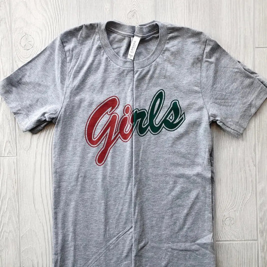 A gray Girls • Tees shirt with Girls in cursive—Gir in red, ls in green—displayed on a light wood floor. Great for matching outfits or Friends-inspired styles.